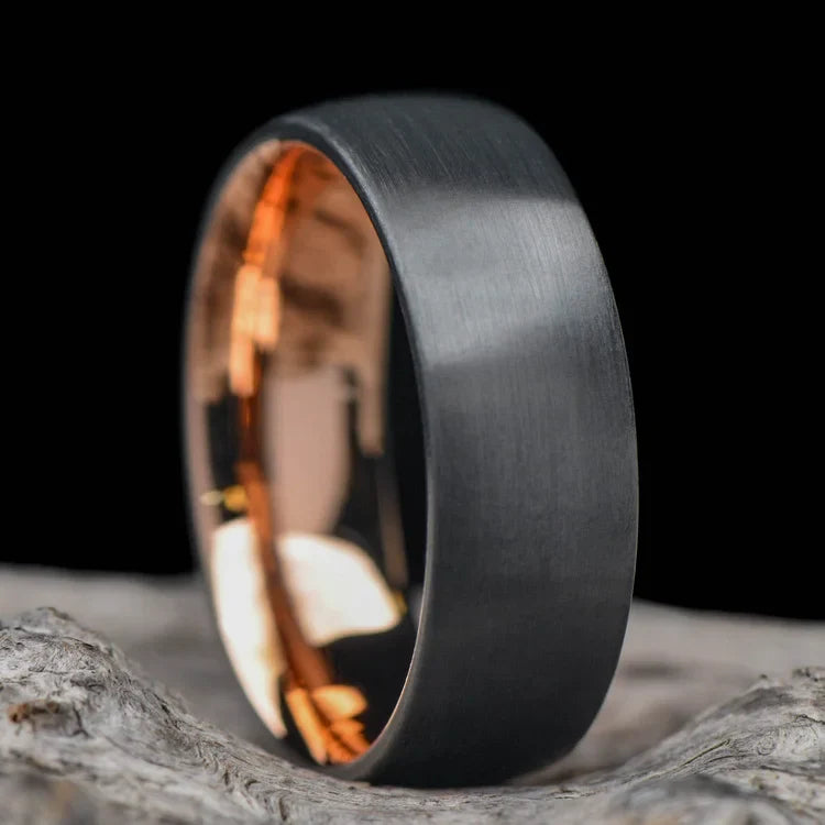 Black titanium wedding band with 18K rose gold inner sleeve, featuring a brushed domed profile, 7mm mens ring, on a white sea branch, free engraving available.