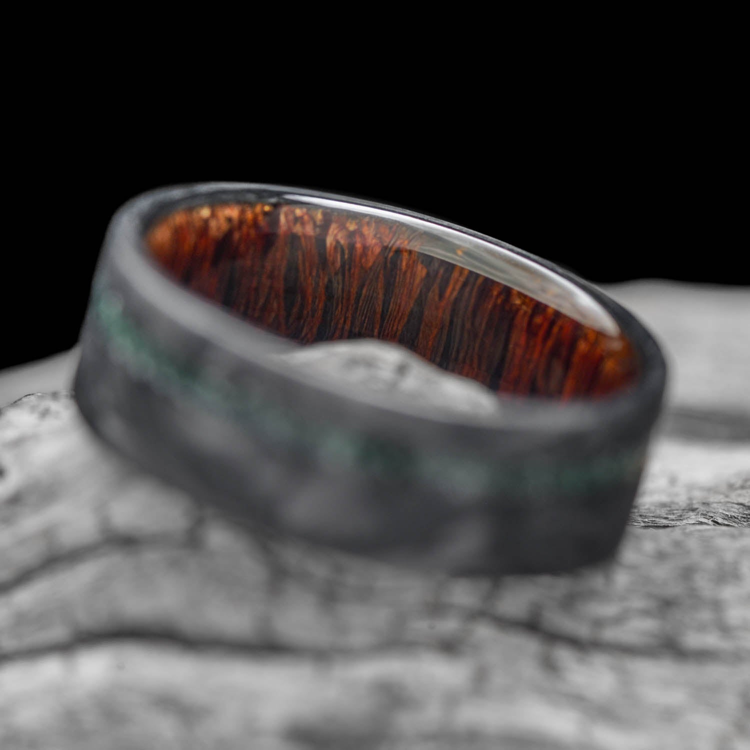 A men's 8mm forged carbon fiber wedding band featuring a vivid green moss agate inlay and a warm leopardwood inner sleeve, offering free engraving, displayed on a white sea branch.