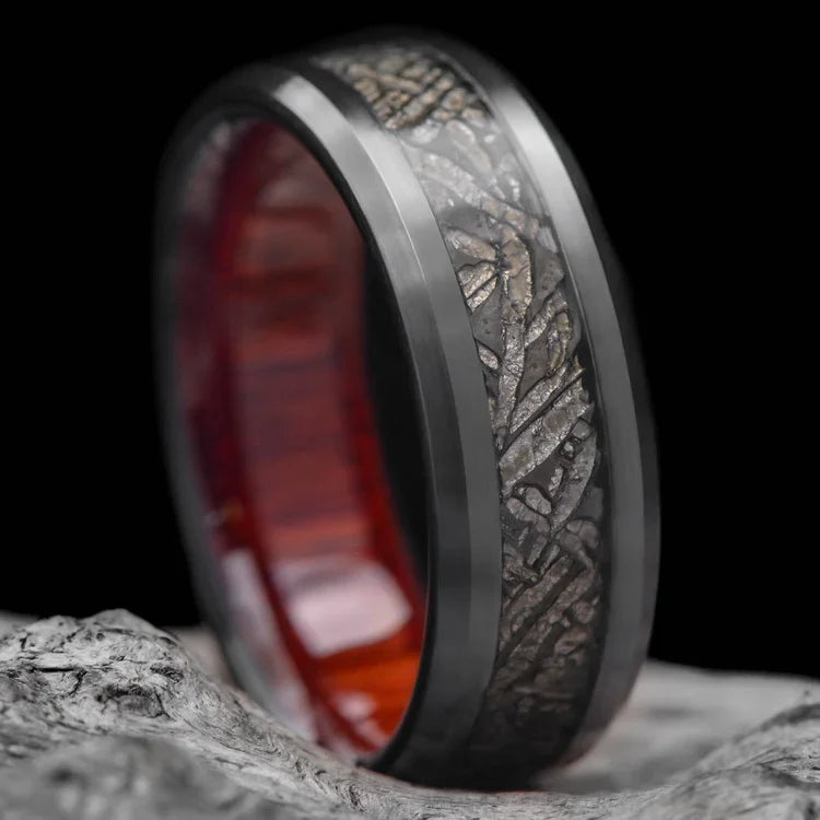 Black titanium ring with central meteorite inlay and bloodwood inner sleeve, 8mm mens wedding band, unique design, free engraving, displayed on a white sea branch.