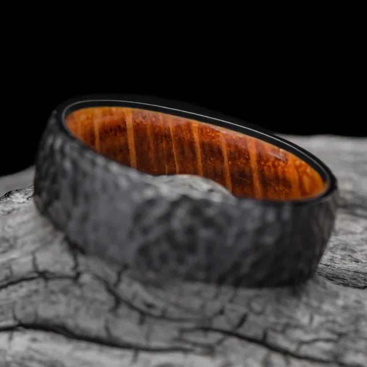 Black titanium wedding band with whiskey barrel oak inner sleeve, featuring a hammered domed design, 7mm mens ring, displayed on a white sea branch, offering free engraving, unique mens wedding band.