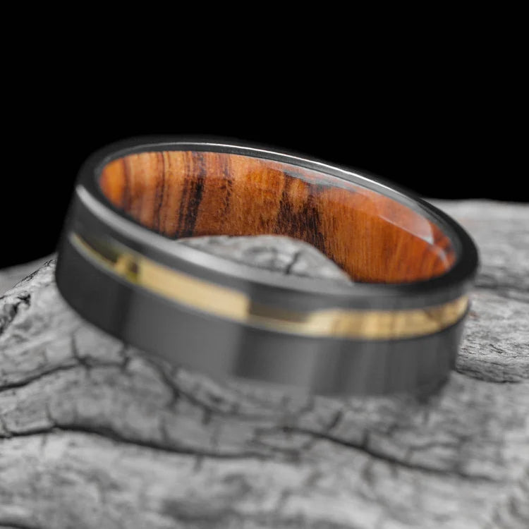 Mens 7mm black titanium wedding band with 18K yellow gold inlay and olivewood inner sleeve, brushed flat profile, free engraving option, on a white background.