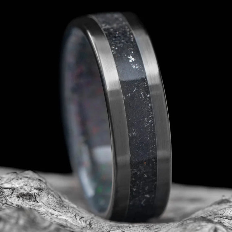 Black Titanium Wedding Band with Meteorite and Sand Inlay, 7mm Mens Ring, featuring a Black Fire Opal Inner Sleeve, displayed on a white sea branch. Free engraving available.