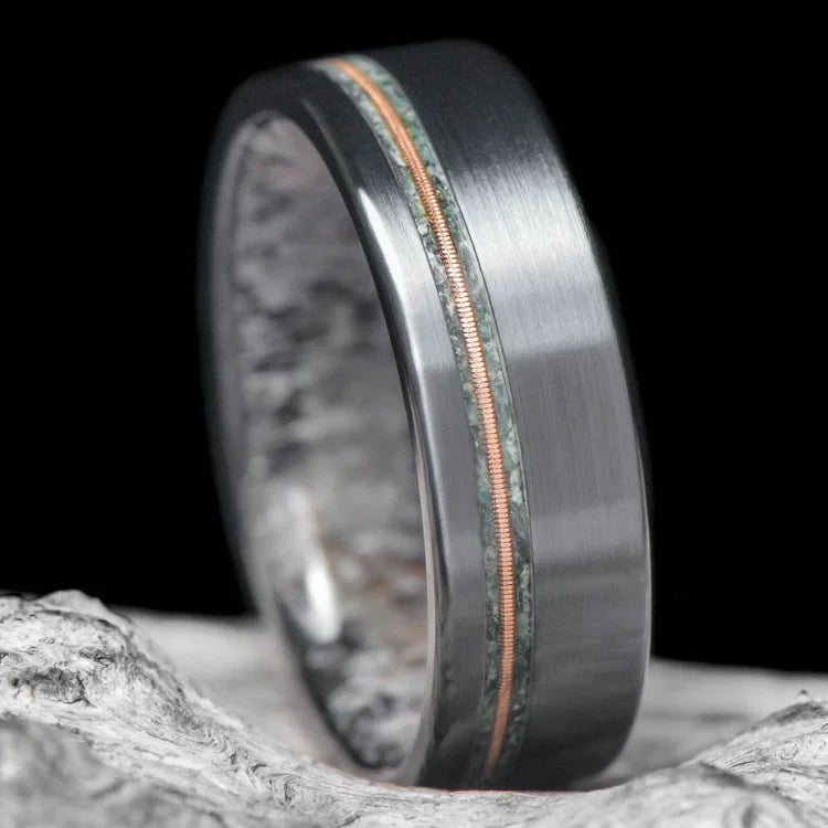 Black titanium wedding band with moss agate and guitar string inlays, featuring free engraving, 7mm width, mens ring with deer antler inner sleeve, displayed on a white sea branch.