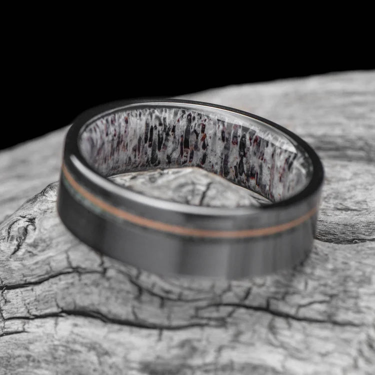 Black titanium mens wedding band with moss agate and guitar string inlay, featuring a deer antler inner sleeve, displayed on a white sea branch, 7mm unique design, free engraving available.