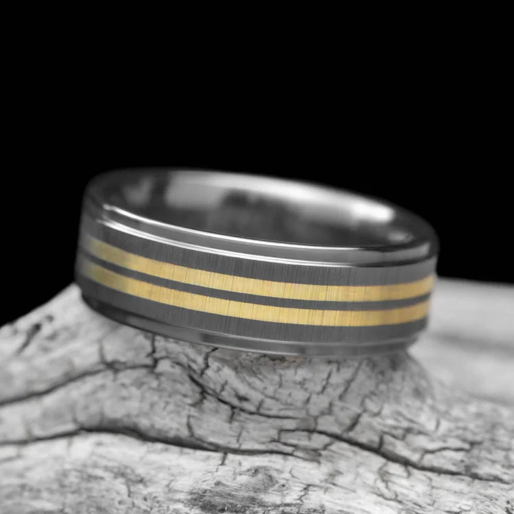 Black titanium wedding band with dual 18K yellow gold inlays, featuring a brushed step profile, displayed on a white sea branch; 7mm mens custom ring with free engraving option.