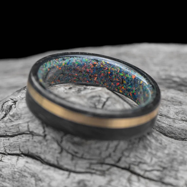Mens 6mm forged wedding band with 14K yellow gold and black fire opal inlay, brushed flat profile, free engraving, displayed on a white sea branch.