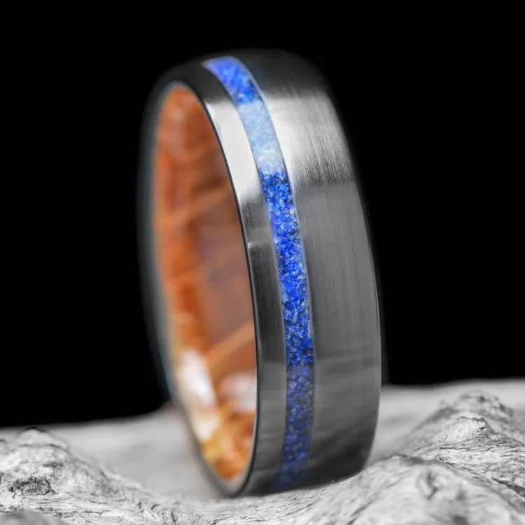 Black titanium wedding ring with lapis lazuli inlay and whiskey barrel oak sleeve, 7mm mens band, on white sand beach.