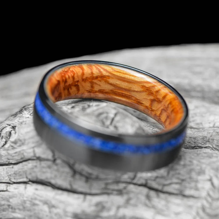 Black titanium ring with lapis lazuli inlay and whiskey barrel oak sleeve, 7mm mens wedding band, unique design, free engraving, displayed on a white sea branch.