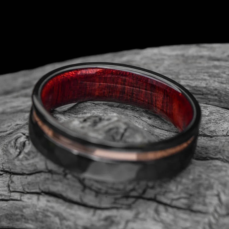 Black titanium mens wedding band with 14K rose gold inlay and bloodwood inner sleeve, faceted flat profile, 7mm, free engraving, displayed on a white sea branch.
