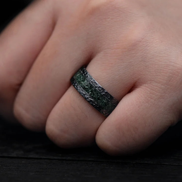 Hand wearing an 8mm black titanium mens wedding band featuring a unique hammered flat profile and central moss agate inlay, offering free engraving.