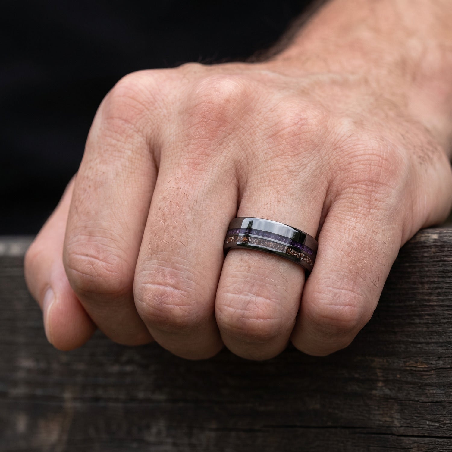 Hand wearing a 6mm black titanium men’s wedding band with crushed T-Rex and amethyst inlays and a black fire opal inner sleeve.