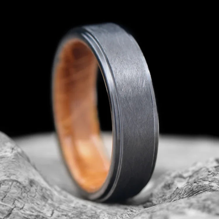 Black titanium wedding band with whiskey barrel oak inner sleeve, featuring a brushed stepped profile, 6mm mens wedding ring, displayed on a white sea branch, available with free engraving.