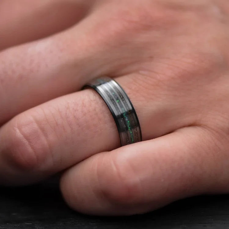 Mens 7mm black titanium wedding band with meteorite and crushed T-Rex inlays, featuring a central black emerald opal, shown on a hand; unique ring with free engraving option.