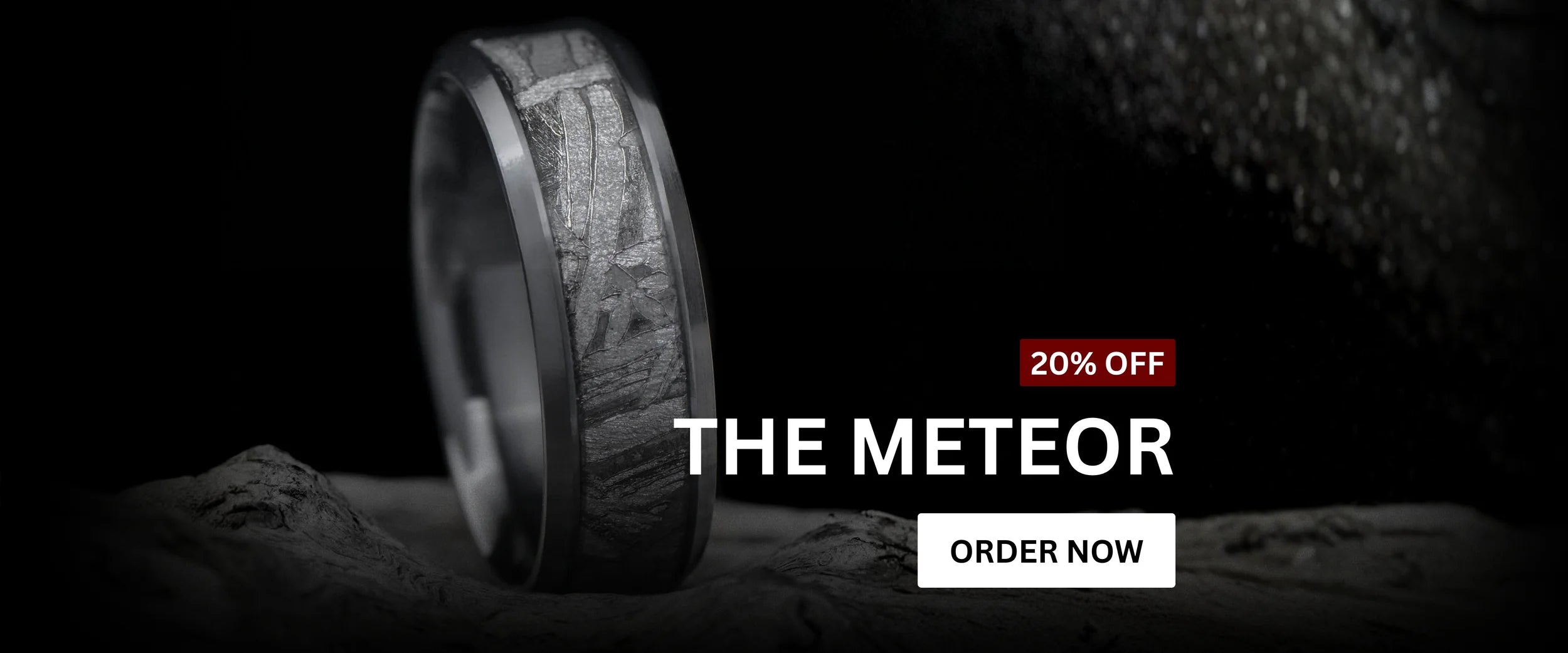 Meteorite wedding ring from The Meteor collection displayed on a textured rock surface. The ring features a dark band with a natural meteorite inlay pattern. Text overlay reads ‘20% Off – The Meteor’ with an ‘Order Now’ button